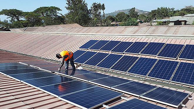 Ein Arbeiter in einer orangefarbenen Sicherheitsweste inspiziert Solarmodule, die auf einem großen Dach installiert sind. Im Hintergrund sind Bäume und Gebäude zu sehen.