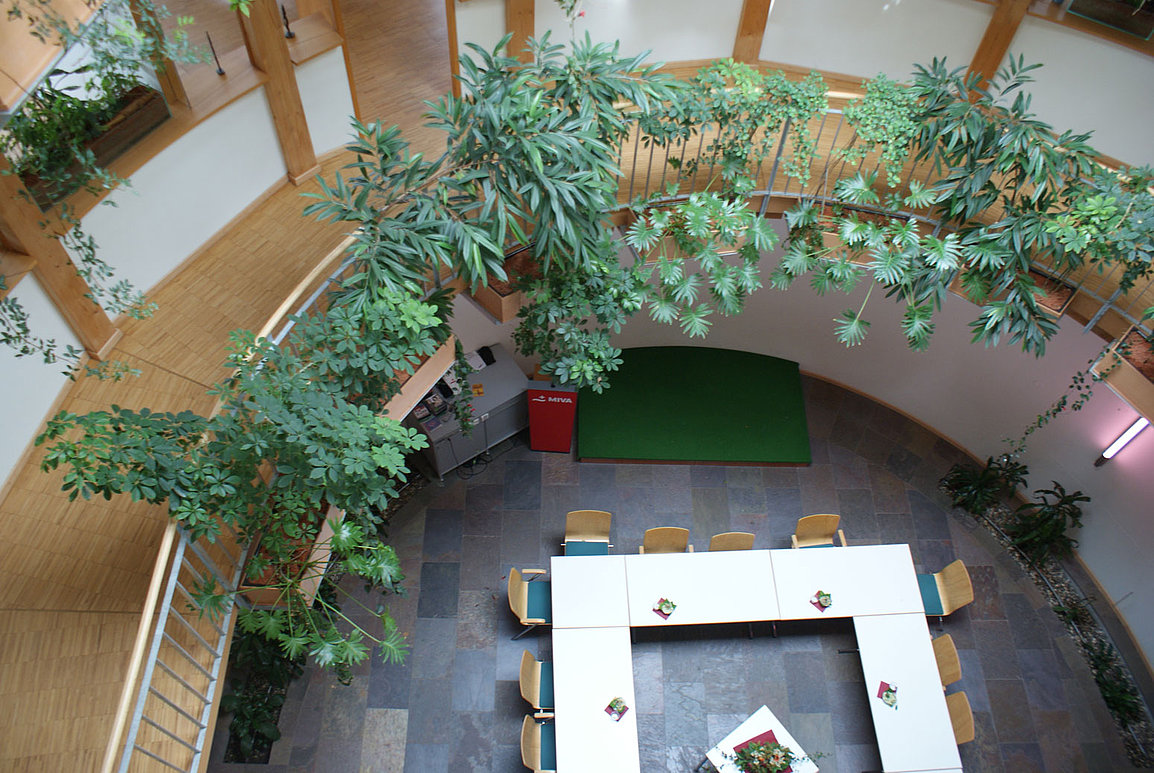 Blick von oben auf ein modernes Innenatrium mit geschwungener Holzbalustrade, üppigem Grün, weißen Tischen in U-Form und hellen Holzstühlen auf einem Fliesenboden..
