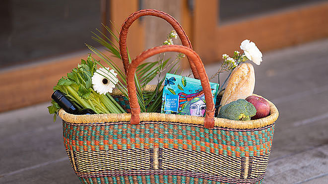 Ein geflochtener Korb mit Ledergriff, gefüllt mit Sellerie, Blumen, verpackten Waren, einem Baguette, Brokkoli und zwei Äpfeln, platziert auf einer Holzoberfläche.