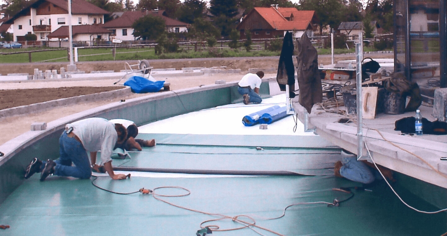 Vier Personen knien und arbeiten an der Installation einer grün-weißen Poolfolie in einem leeren Wasserbecken.