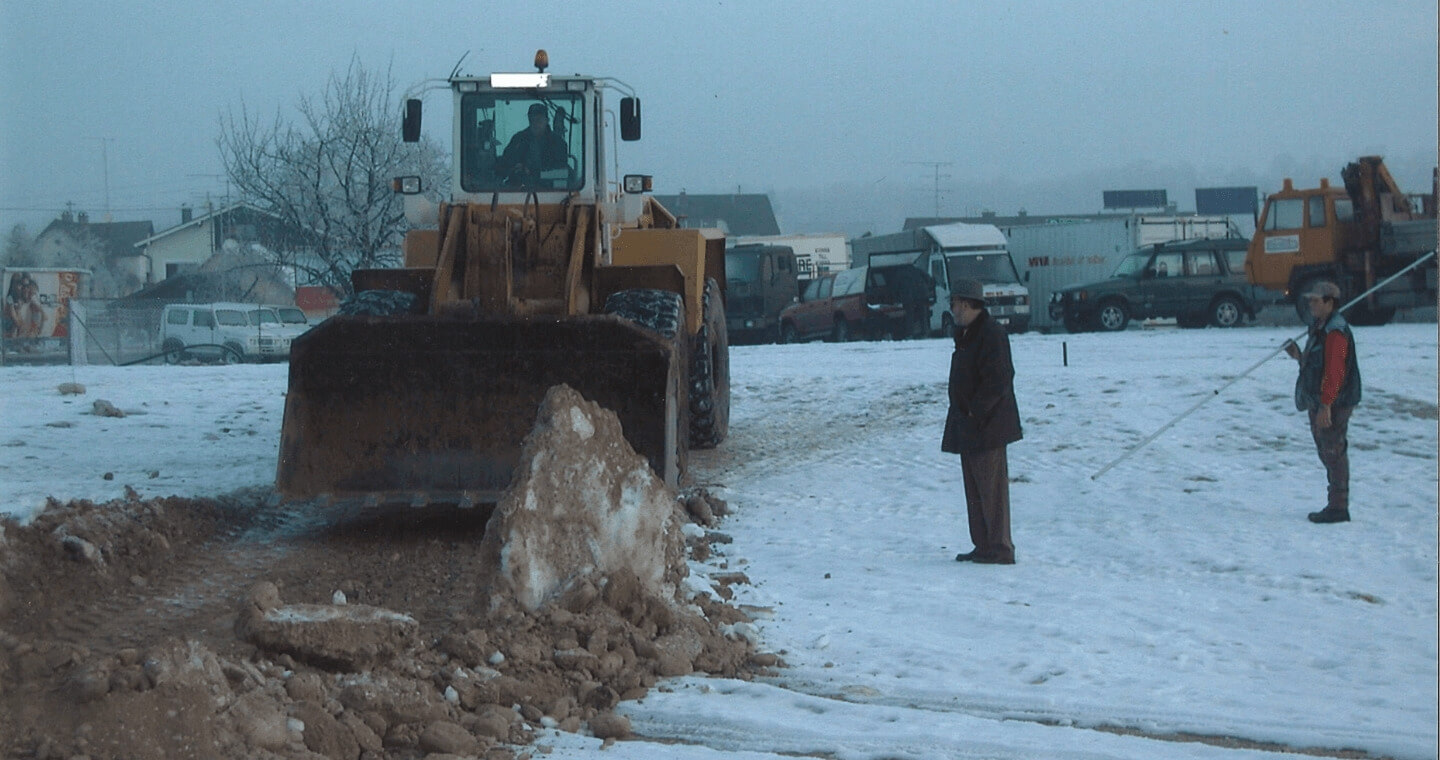 Ein großer gelber Bulldozer bewegt Erde und Schnee auf einer verschneiten Baustelle. Zwei Personen stehen in der Nähe. Eine beobachtet die Baustelle aufmerksam, die andere hält weiter entfernt eine Stange.