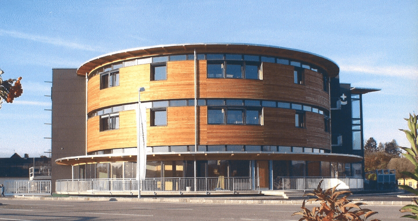 Ein modernes, dreistöckiges Rundgebäude mit Holzvertäfelung und großen Glasfenstern vor einem strahlend blauen Himmel. Das Gebäude ist im Erdgeschoss von einem Geländer umgeben und liegt an einer Straße.