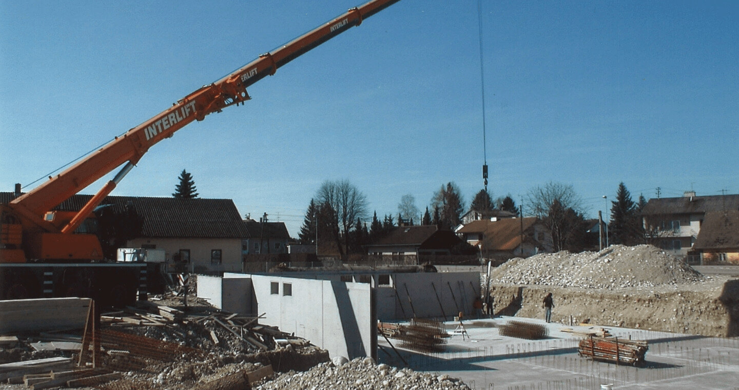 Ein orangefarbener Kran hebt Materialien auf einer Baustelle mit Betonwänden, Baumaterialien und Kieshaufen. 