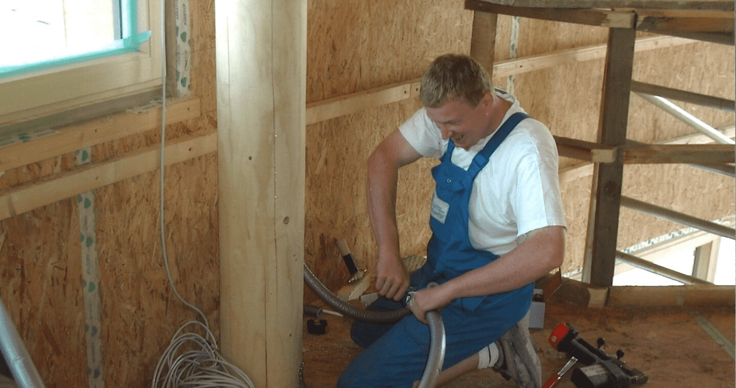 Ein Arbeiter im blauen Overall installiert elektrische Leitungen in einem im Bau befindlichen Raum mit Holzrahmen. Er kniet auf dem Boden und benutzt Werkzeuge in der Nähe eines Fensters.