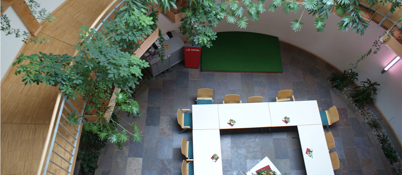 Blick von einem Stockwerk nach unten auf einen modernen Besprechungsraum mit weißen, U-förmig angeordneten Tischen, umgeben von Stühlen. Von oben hängen grüne Pflanzen herab.