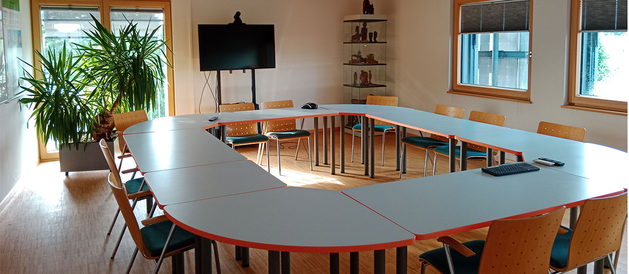 Ein heller Seminarraum mit U-förmig angeordneten Tischen, Holzstühlen, einer großen Topfpflanze, einem Fernsehbildschirm, dekorierten Ausstellungsregalen und Fenstern, die natürliches Licht hereinlassen.