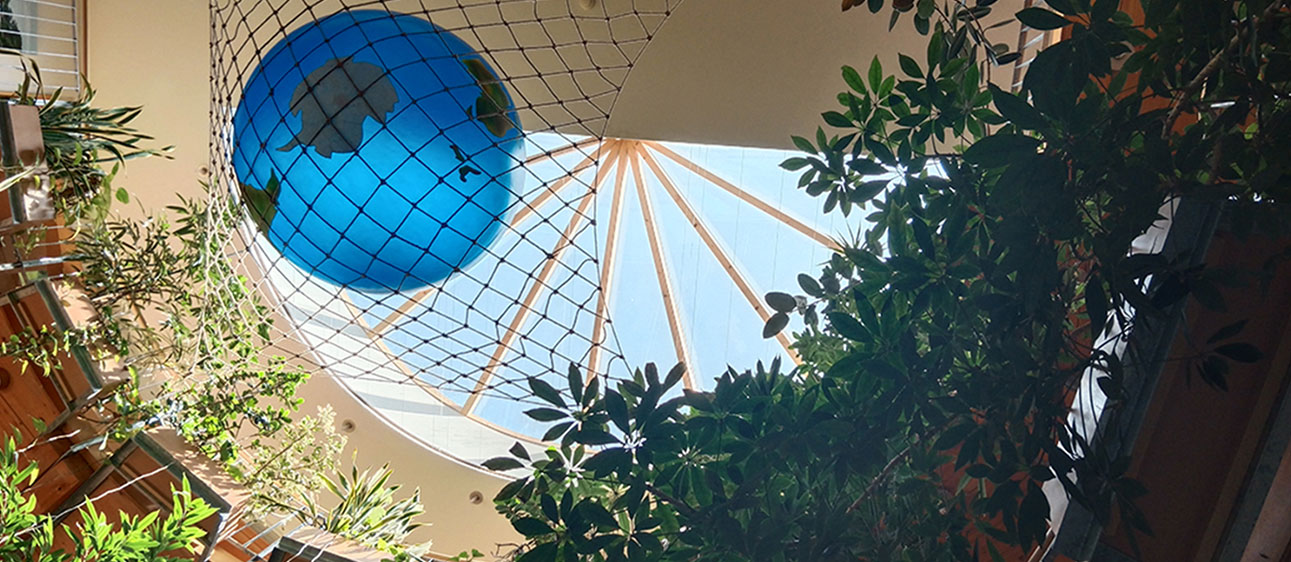 ine große Weltkugel liegt in einem Netz, das von der Decke eines Atriums hängt. Das Atrium ist von grünen Zimmerpflanzen und runden Balkonen umgeben, und natürliches Licht fällt von oben ein.