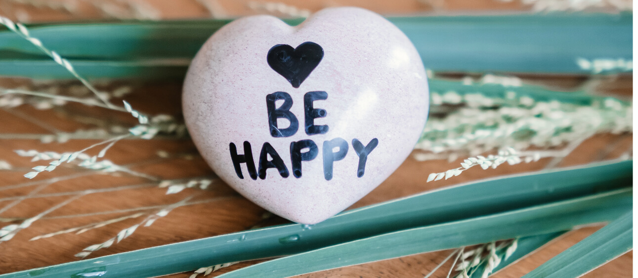 Ein rosafarbener herzförmiger Stein mit einem schwarzen Herzsymbol und den Worten „Be Happy“ liegt zwischen grünen Blättern und getrockneten Gräsern auf einer Holzoberfläche.