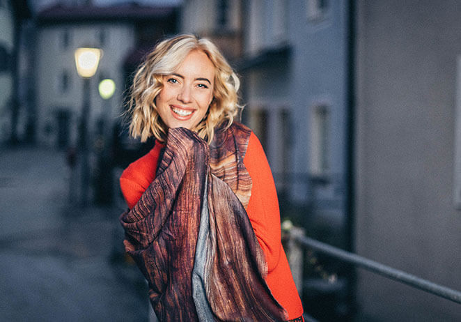 Eine lächelnde Frau mit kurzen blonden Haaren steht draußen auf einer Straße. Sie trägt einen roten Pullover und hält einen gemusterten Schal vor ihr Gesicht. Der Hintergrund ist durch Gebäude und eine Straßenlaterne leicht verschwommen.