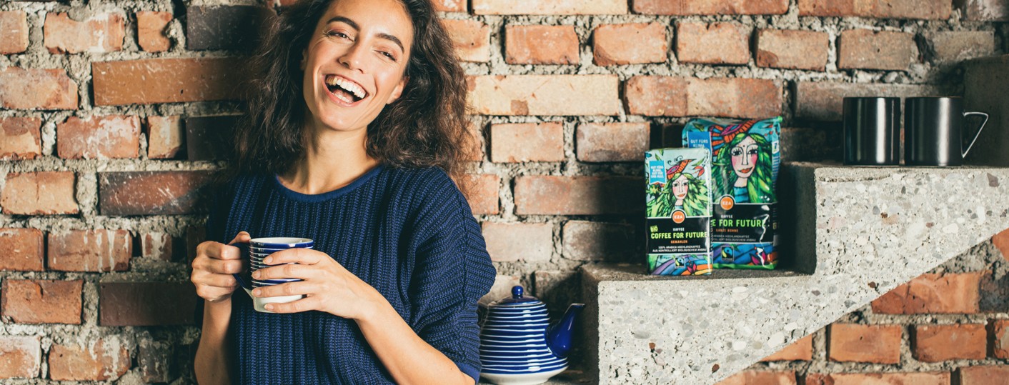 Eine lächelnde Frau in einem marineblauen Pullover hält einen blau gestreiften Becher und steht neben einer Backsteinmauer und Steinstufen, auf denen bunte Kaffeeverpackungen, eine blaue Teekanne und Becher ausgestellt sind.